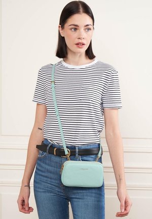 Sac en cuir bleu clair à bandoulière avec des accessoires en or, de forme rectangulaire et avec une sangle ajustable, porté sur une chemise rayée et un jean bleu.