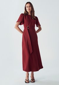 Robe midi rouge avec des manches courtes bouffantes et une ceinture à la taille ornée d'une boucle en or. Tissu lisse, design fluide et texture locale.