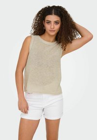 Beige mouwloze gebreide top met een ronde halslijn en subtiele textuur, gecombineerd met opgerolde witte denim shorts. Neutrale kleuren creëren een lichte, casual uitstraling.