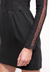 Vestido negro con un Cuerpo de encaje y largas mangas transparentes con lunares. Falda de satén suave con una silueta ajustada y un ligero brillo.