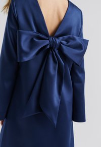 Robe satin marine avec un profond décolleté en V dans le dos et un grand nœud à l'arrière. Manches longues avec une texture lisse et brillante.