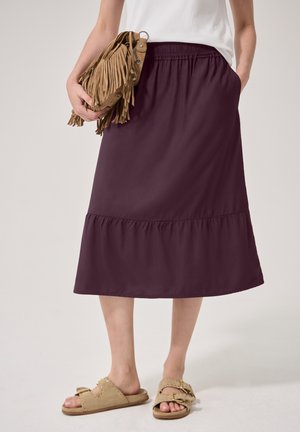 Donna con gonna midi viola scuro, sandali beige, top bianco e che tiene una borsa a frange in suede color cammello al suo fianco.