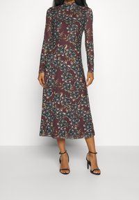 Robe à manches longues, bordeaux, avec un motif floral dans des teintes de vert, bleu et jaune. Silhouette fluide, longueur cheville, associée à des talons noirs à lanières.