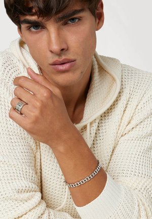 Bracelet en argent avec des maillons géométriques imbriqués, porté sur une main. La personne porte un hoodie en tricot crème à texture.