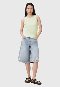 Top sin mangas de color verde claro, pantalones cortos de mezclilla azul claro de corte holgado con un parche desgastado y sandalias negras con acentos dorados.