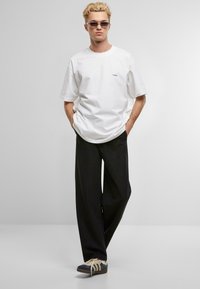Hvit bomull oversized t-skjorte med en liten blå logo, kombinert med svarte brede bukser og mørke joggesko med kremfargede detaljer.