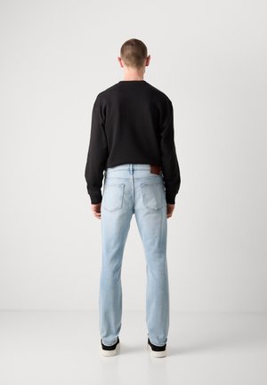 Mann steht mit dem Rücken zur Kamera, trägt ein schwarzes Langarmshirt, hellblaue Jeans mit Gesäßtaschen und schwarz-weiße Turnschuhe.