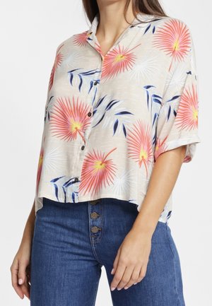 Vrouw draagt een beige korte mouwen blouse met knopen en koraal- en witte bloemenprints, gecombineerd met een hoog geplaatste blauwe spijkerbroek.