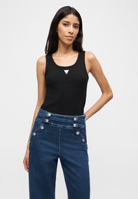 Γυναίκα με μακριά σκούρα μαλλιά, φορώντας μαύρο αμάνικο τοπ και ψηλόμεσο μπλε τζιν με ασημένια κουμπιά, που ποζάρει μπροστά σε ουδέτερο φόντο.