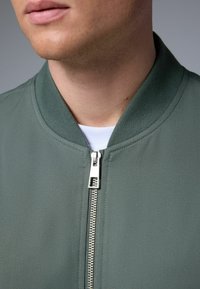 Zelena bomber jakna s rebrastim ovratnikom, koja ima srebrni patent zatvarač i teksturiranu tkaninu. Približeni pogled usredotočen na područje dekoltea.