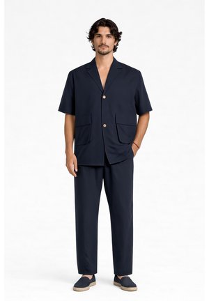 Uomo con capelli corti e scuri che indossa un blazer blu navy a maniche corte, pantaloni abbinati e espadrillas slip-on, in piedi con una mano in tasca.