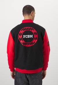 Giacca varsity nera e rossa con colletto a costine. Presenta una grande grafica circolare con "FCBM" e un motivo a rombi sul retro.