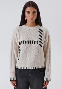 Maglione lavorato a maglia spesso con blocchi di colore crema e nero. Presenta motivi a treccia e dettagli con frange. Orlo e polsini a costine.