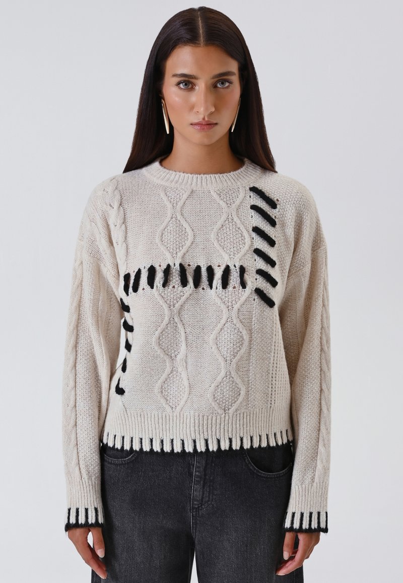 Maglione lavorato a maglia spesso con blocchi di colore crema e nero. Presenta motivi a treccia e dettagli con frange. Orlo e polsini a costine.
