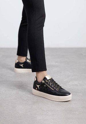 Zapatillas de cuero negro con detalles en dorado, textura acolchada y suela blanca. Presentan cordones y una cremallera decorativa.