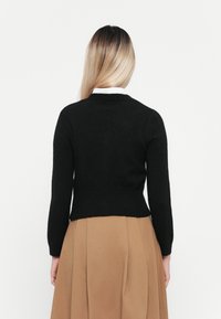 Person with long blonde hair wearing a black sweater over a white shirt and a brown pleated skirt, facing away on a white background.