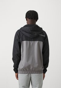 Huvtröja med en svart övre del, ljusgrå nedre del, elastiska ärmslut och en zipficka på den övre ryggen. Slät tygtextur.