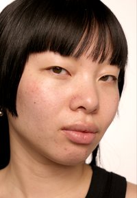 Portrett av en person med kort svart hår og rettlugg, jevn hud med noen blemmer, og lipper i nøytrale farger.