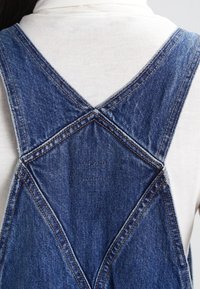 Denim overalls feature a crisscross back design, with contrast stitching and a faded blue color, highlighting the fabric's texture and durability.