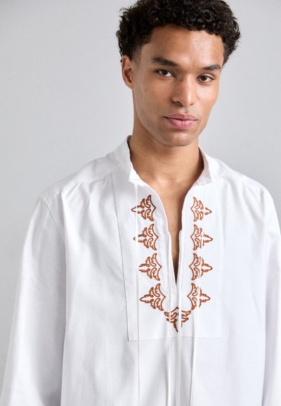 Tunique blanche à décolleté en V profond, ornée de broderies marron délicates le long de l'encolure. Manches longues, texture lisse et tissu léger.