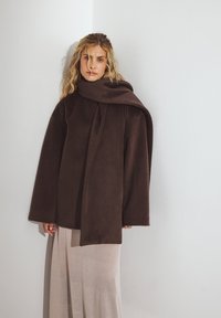 Üks suurte mahuga pruun flanellmantel kõrge kraega ja lipsuga detailiga, kantud heledat tooni voolava seeliku üle. Sile tekstuur, minimaalne disain.