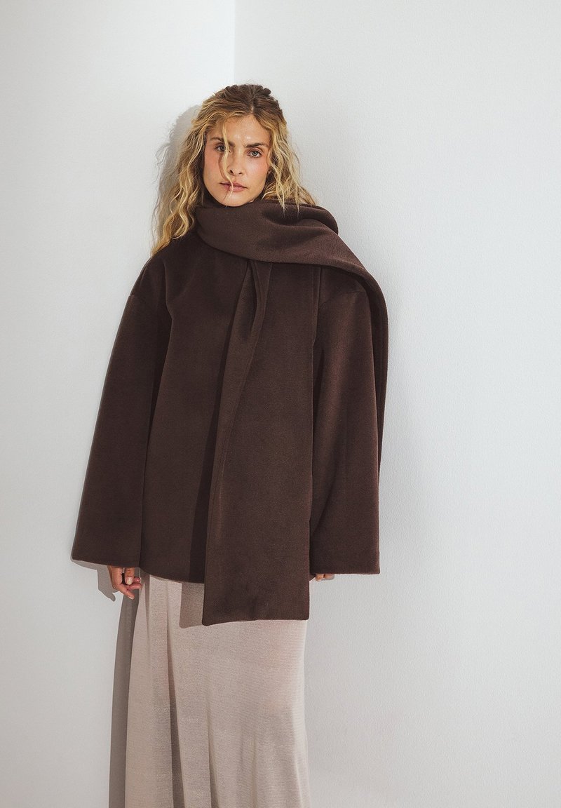 Üks suurte mahuga pruun flanellmantel kõrge kraega ja lipsuga detailiga, kantud heledat tooni voolava seeliku üle. Sile tekstuur, minimaalne disain.