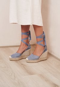 Espadrillas a zeppa in camoscio blu con laccetti alla caviglia, suola in giunco naturale e punta intrecciata bianca. Superficie strutturata e design liscio.