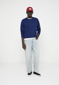 Pull bleu avec le texte en relief "Originals", t-shirt blanc en dessous, jeans clairs amples et baskets noires. Une casquette rouge vient compléter la tenue.