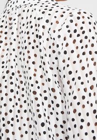 Blusa bianca trasparente con un motivo di pois neri e marroni, con strisce orizzontali nel tessuto e una sottile trama.