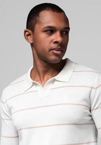 Muž s krátkými vlasy v bílém košelovém triku s tenkými béžovými vodorovnými pruhy, dívající se do strany na jednobarevném šedém pozadí.