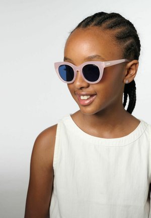 Jeune fille aux cheveux tressés portant des lunettes de soleil rose clair et un débardeur blanc, souriante et regardant vers la droite.