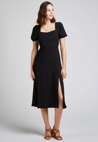 Robe midi noire avec décolleté carré et manches bouffantes. Présente un tissu texturé et une fente sur le côté. Associée à des sandales à lanières brunes.