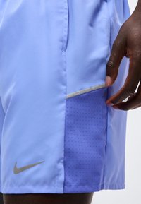 Κοντινή λήψη ανοιχτό μπλε αθλητικού σόρτς με διάτρητο πλαϊνό πάνελ και ένα χέρι που φτάνει προς την τσέπη, με αντανακλαστικό λογότυπο Nike Swoosh.