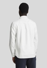 Witte blouse met lange mouwen en een button-down kraag, met een yoke en zijsplitten aan de zoom. Het materiaal lijkt zacht en soepel.
