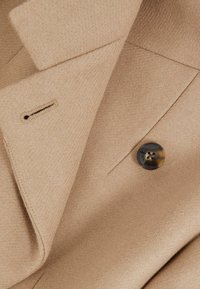 Beige räätälöity takki, jossa on näkyvä nappilenkki ja tumma ruskea neljän reiän nappi tekstuurilla varustetussa kankaassa.