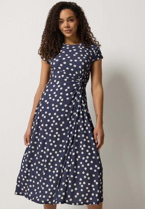 Vrouw met krullend haar, gekleed in een marineblauwe jurk met witte polkadots, voorzien van een zijdetail met twist en korte mouwen.