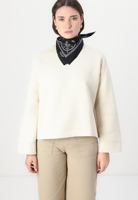 Felpa color crema con vestibilità rilassata, dotata di maniche lunghe e scollatura a V. Bandana nera con motivo bianco annodata attorno al collo.
