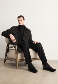 Čierny trenčkot cez čierny rolák, skombinovaný s čiernymi nohavicami a topánkami. Model sediaci na béžovej stoličke, minimalistické pozadie.