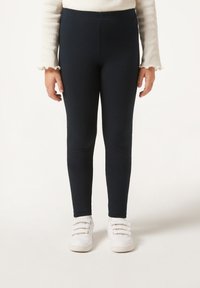 Marineblaue Leggings aus einem dehnbaren Stoff, knöchellang, kombiniert mit weißen Sneakers, die mehrere Schnürsenkelöffnungen und klare Linien aufweisen.