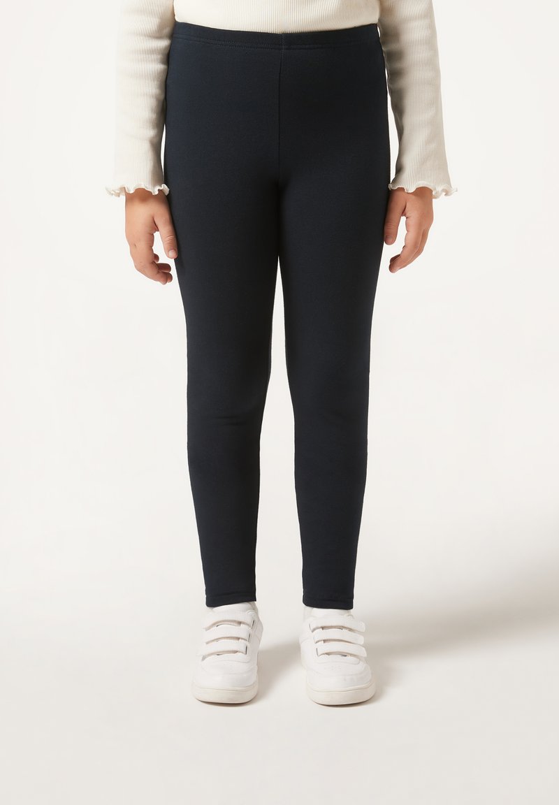 Marineblaue Leggings aus einem dehnbaren Stoff, knöchellang, kombiniert mit weißen Sneakers, die mehrere Schnürsenkelöffnungen und klare Linien aufweisen.