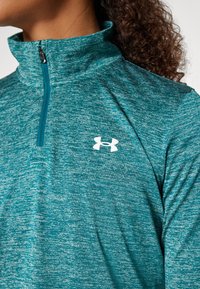Teal, halvzip träningsjacka i strukturerad tyg med en vit logotyp på bröstet. Hög krage och slät dragkedja.