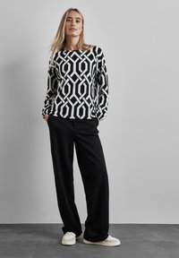 Schwarz-weißes Langarmshirt mit geometrischem Muster, kombiniert mit lockeren schwarzen Hosen und hellfarbenen Schuhen.