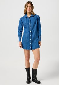 Denim haljina košulja u plavoj boji sa dugmićima, dugim rukavima, džepovima na grudima i šiljastim detaljima na yoku. Uparena s crnim čizmama do koljena.