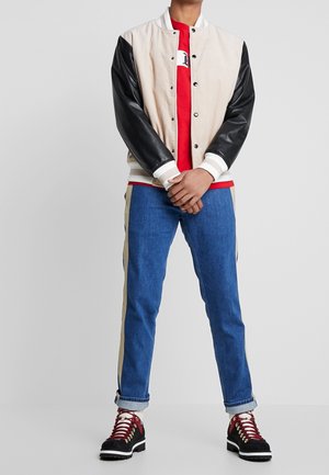 Chaqueta bomber de pana beige y cuero negro, camiseta roja, vaqueros azules con franjas laterales beige, y botas de senderismo negras con cordones rojos.