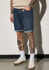 Shorts de mezclilla, lavado oscuro, con dobladillo enrollado, combinados con zapatillas blancas y calcetas acanaladas. Tatuajes en las piernas notables visibles. Fondo de suelo de concreto.
