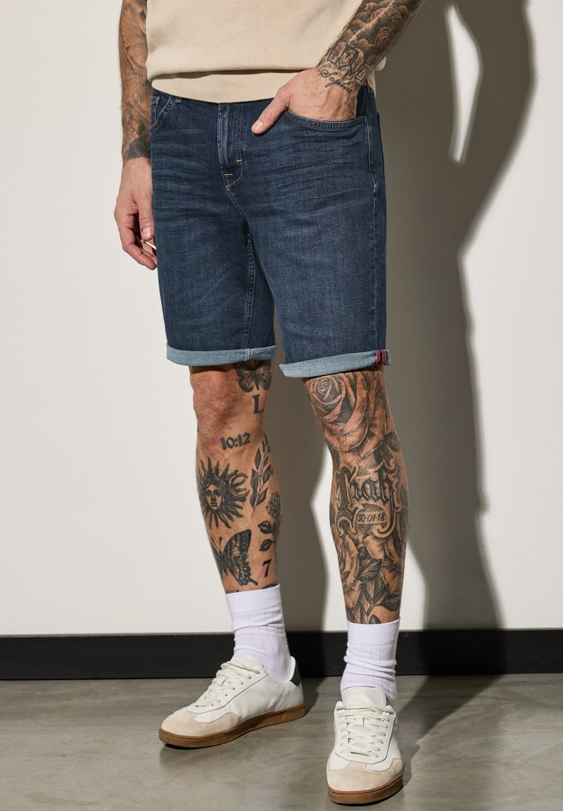 Shorts de mezclilla, lavado oscuro, con dobladillo enrollado, combinados con zapatillas blancas y calcetas acanaladas. Tatuajes en las piernas notables visibles. Fondo de suelo de concreto.