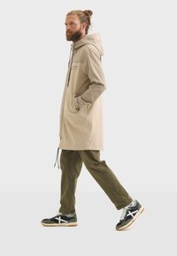 Un homme avec une barbe marche dans un manteau beige, un pantalon vert olive et des baskets à la mode sur un fond blanc uni.