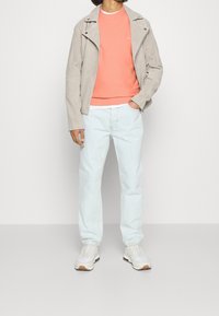 Hellgraue Wildleder-Motorradjacke über einem korallenfarbenen Sweatshirt, kombiniert mit hellblauen Jeans und weißen Sneakers mit sandfarbenen Akzenten.