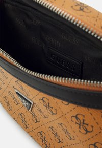 Bruine handtas met een patroon, zwarte leren accenten en een zilveren rits, voorzien van een merktag aan de binnenkant en een getextureerde buitenkant.