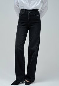 Persona que lleva unos pantalones rectos de talle alto de color negro, una camisa blanca abotonada metida por dentro y unos zapatos de tacón negro con punta puntiaguda, de pie contra un fondo gris.